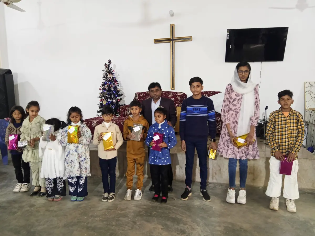 Children standing in a row holding gift parcels beneath a wooden cross in a church hall
